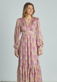 Langes Kleid mit Puffärmeln aus lila und gelb gemustertem Stoff. V-Ausschnitt mit tailliertem Bund und gestuften Rock. Sanfte Textur, fließende Silhouette.