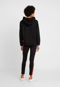 Sudadera negra con capucha y mangas largas, combinada con joggers negros que tienen franjas laterales rojas y zapatillas blancas. Tejido suave, corte casual.