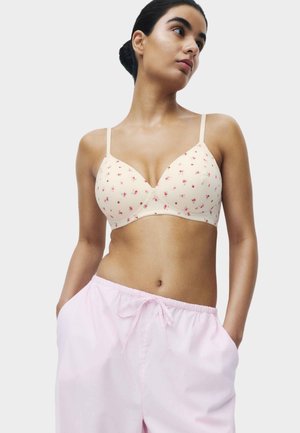 Frau trägt einen cremefarbenen BH mit kleinen rosa Blumenmustern und hellrosa Hose mit Kordelzug, steht mit den Händen in den Taschen und schaut zur Seite.