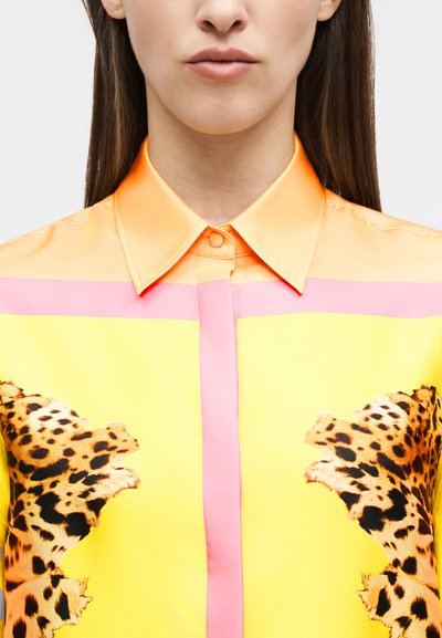 Γυναίκα που φοράει φωτεινό κίτρινο πουκάμισο με ροζ ρίγες, πο ρτοκαλί γιακά και μοτίβα λεοπάρ στο στήθος.