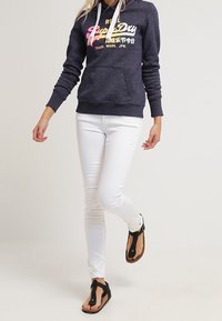 Sudadera marina con logo blanco y rosa, bolsillo frontal; jeans slim-fit blancos; chanclas negras con tira en el dedo y suela plana.