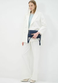 Witte op maat gemaakte blazer en wijde broekset, gecombineerd met een marineblauwe leren tas met contrasterende stiksels en een lange verstelbare schouderriem.
