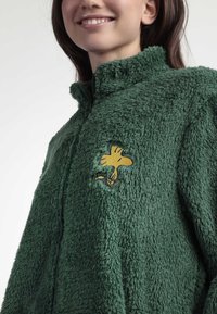Mujer sonriendo, vestida con una chaqueta de felpa con cierre en color verde oscuro y un pequeño pájaro de dibujos animados amarillo bordado en el pecho.