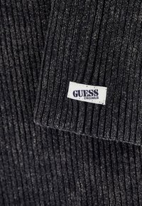 Tissu tricot côtelé foncé avec une petite étiquette rectangulaire blanche cousue, affichant "GUESS ORIGINALS" en lettres noires.
