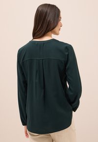 Dunkelgrüne Bluse mit langen Ärmeln, rundem Ausschnitt und einem gefältelten Rückendetail. Glatte Textur und lockere Passform mit leicht abgerundetem Saum.