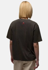 Personne avec une coupe de cheveux bob blonde portant un t-shirt noir avec un petit logo Jumpman rouge dans le haut du dos, et un pantalon beige.