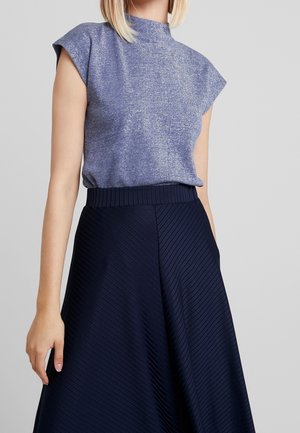 Femme portant un haut bleu brillant sans manches, rentré dans une jupe plissée taille haute bleu marine foncé, sur un fond gris uni.