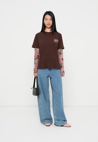 Brown cotton t-shirt with short sleeves and logo, layered over long-sleeved patterned shirt, paired with light blue wide-leg jeans and a small black bag.