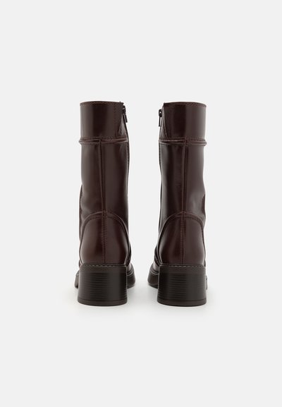 Brūni ādas potītes zābaki ar gludu tekstūru, taisnstūra papēdi un sānu rāvējslēdzēju. Ievērojamas šūšanas detaļas gar šuvēm.