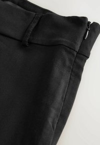 Gros plan sur un pantalon noir avec passant de ceinture et fermeture éclair latérale sur fond clair.