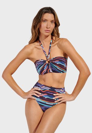 Mujer con bikini halter a rayas de colores y braguitas de talle alto, posando con las manos en las caderas frente a un fondo liso.