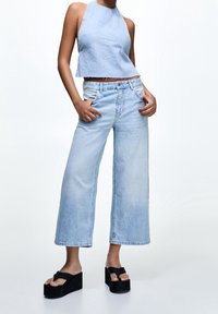 Světle modrý denimový halter top v kombinaci s širokými světle modrými džíny. Modelka má na sobě černé platformové sandály, což vytváří ležérní outfit.