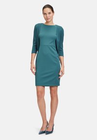 Tealowa sukienka dopasowana z rękawami o długości trzech czwartych i usztywnioną górą. Gładka faktura materiału, długość do kolan, w zestawieniu z granatowymi szpilkami.