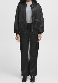 Schwarze Satin-Bomberjacke mit Reißverschluss vorne, Seitentaschen und gerippten Bündchen, kombiniert mit schwarzen, weit geschnittenen Cargo-Hosen und glänzenden Schuhen.