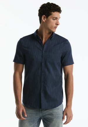 Man met kort krullend haar die een donkerblauw kort mouwen overhemd draagt en lichtblauwe spijkerbroek, kijkt naar zijn rechterkant.