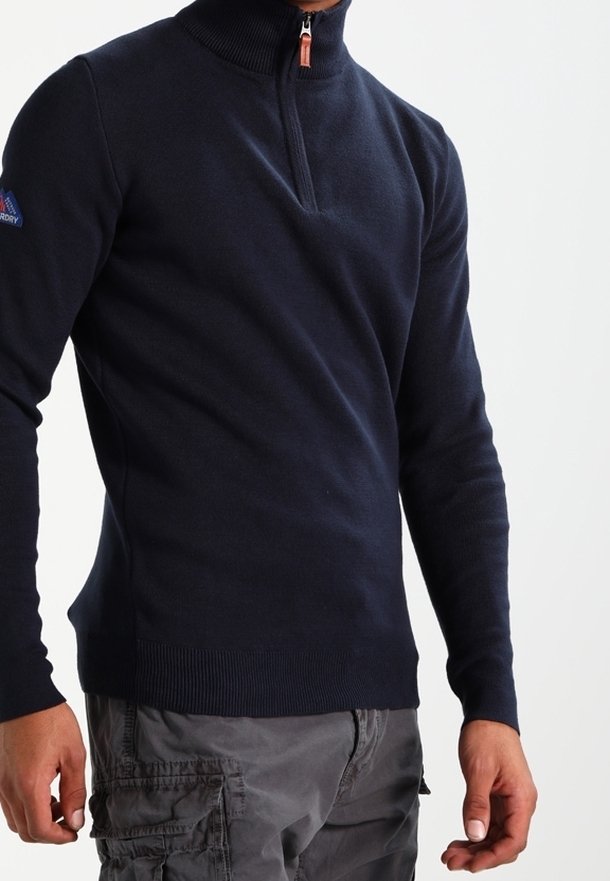 Homme portant un pull à manches longues bleu marine à demi-fermeture éclair et un pantalon cargo gris, debout avec les bras détendus le long du corps.