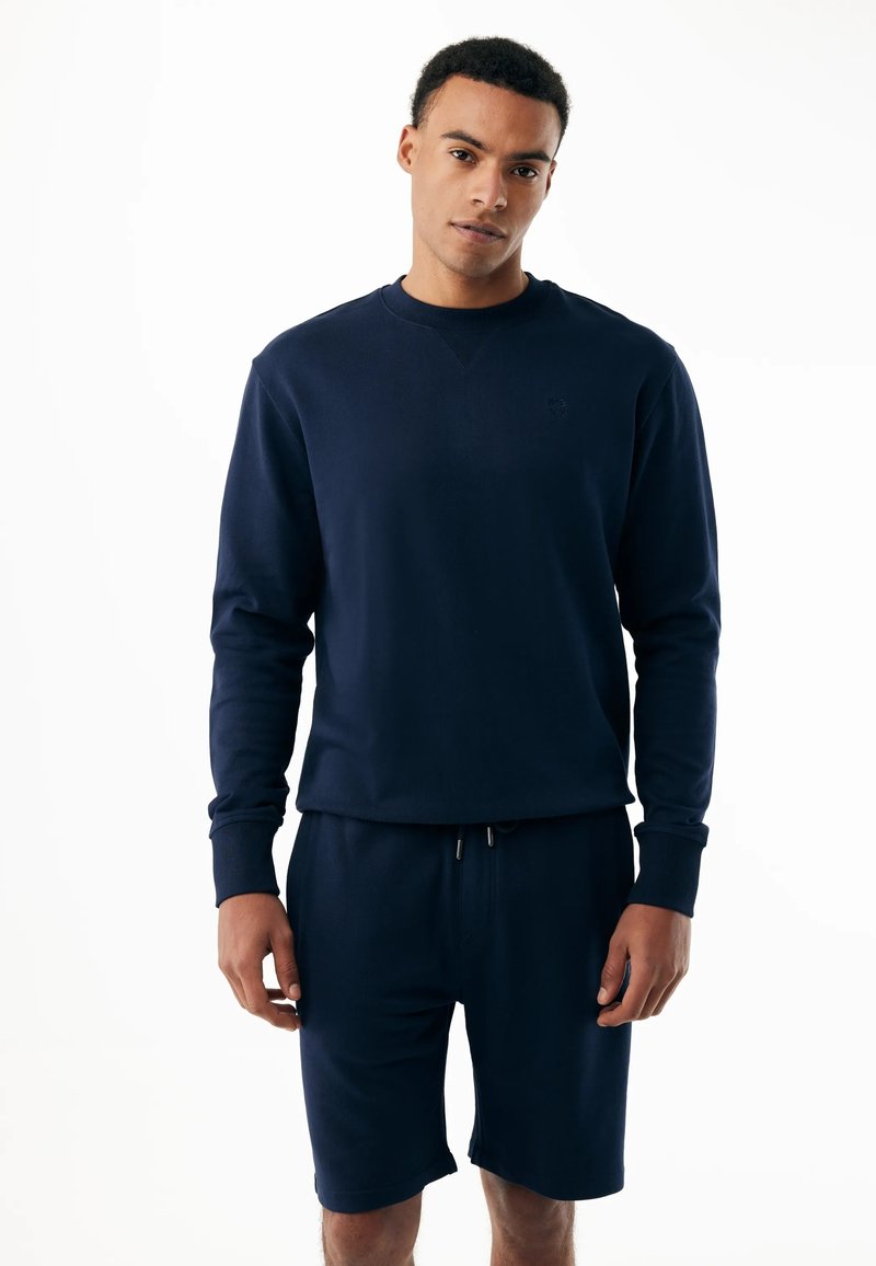 Jonge man draagt een marineblauwe sweater met lange mouwen en bijpassende korte broek, staand tegen een witte achtergrond.
