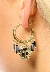 Boucle d'oreille en or avec un design central en forme de scarabée, présentant des accents noirs et des tiges noires et blanches pendantes. Détails de surface texturée visibles.