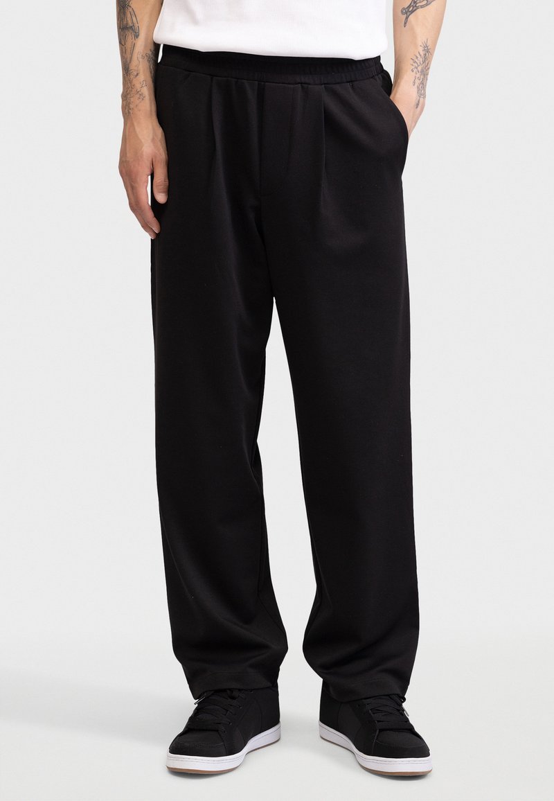 Άνδρας με φαρδιά μαύρα παντελόνια, μαύρα αθλητικά παπούτσια με λευκές σόλες, στέκεται με το ένα χέρι στην τσέπη μπροστά σε απλό φωτεινό φόντο.