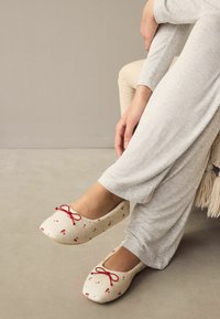 Ballet flats dengan kain krim, corak ceri merah, dan detail busur kecil berwarna merah. Desain ujung bulat yang lembut, dipadukan dengan celana santai abu-abu muda.