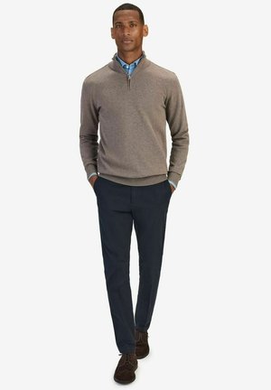 Bruine gebreide trui met een kwart-rits kraag, gecombineerd met marineblauwe broek. Model draagt donkerbruine suède schoenen en een lichtblauw shirt eronder.