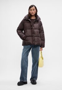 Smeđa pufasta jakna s visokim ovratnikom, s velikim kvadratnim dijelovima i kopčama na gumb. Nosena s svijetloplavim trapericama i žutom torbicom.