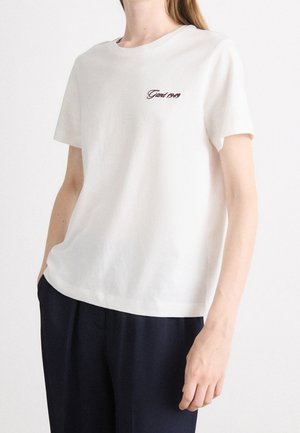 Femme portant un t-shirt blanc uni à manches courtes avec le texte "Gant 1949" sur la poitrine et un pantalon noir sur fond blanc.