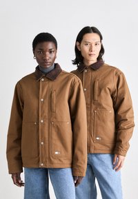 Brune bomuldsjakker med kordfløjlskraver, to forlommer og trykknaplukninger. Casual pasform, båret over lyse turtlenecks og blå jeans.