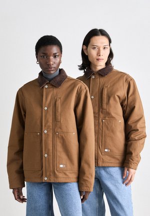Bruine katoenen jassen met corduroy kraag, twee voorzakken en drukknoopsluitingen. Casual pasvorm, gedragen over lichte coltruien en blauwe jeans.