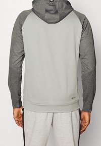 Sweat à capuche gris clair avec manches gris foncé, texture tricotée et ourlet ajusté. Présente un détail de logo dans le bas du dos.