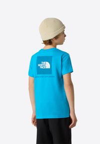 Turquoise children's t-shirt with a crew neck, featuring a centered white logo and text on the back. The fabric appears smooth and lightweight.