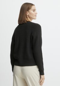 Pull en maille côtelée noir avec manches longues et col rond, comportant un ourlet légèrement court, porté avec un pantalon couleur crème.