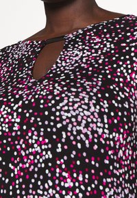 Lähellä otettu kuva tummaihoisesta henkilöstä, joka käyttää mustaa ykköstä, jossa on pinkkejä, valkoisia ja violetteja pilkkuja, sekä pientä avainta muistuttavaa kaula-aukkoa.