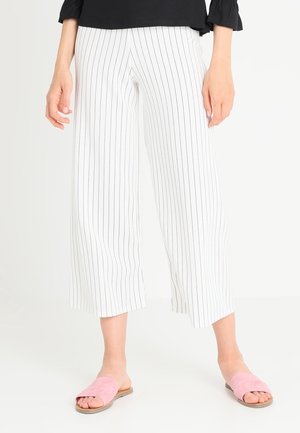 Vrouw met witte wijde cropped broek met zwarte verticale strepen en roze slippers, staand voor een effen achtergrond.