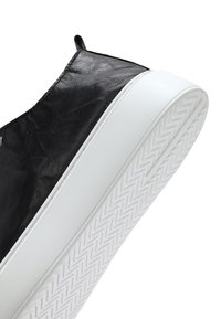 Zwarte leren sneaker met dikke witte rubberen zool, schuin weergegeven met nadruk op het geprofileerde zigzagloopvlak van de zool.