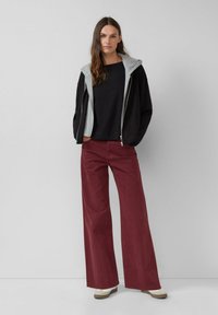 Vrouw staat met handen in de zakken, gekleed in een zwarte trui, een zwart en grijs gevoerd jack, brede bordeauxrode broek en beige schoenen.