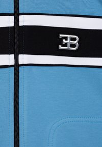 Giacca zip azzurra chiara con strisce orizzontali bianche e nere, accentuata da un logo argento e una texture liscia con dettagli cuciti.