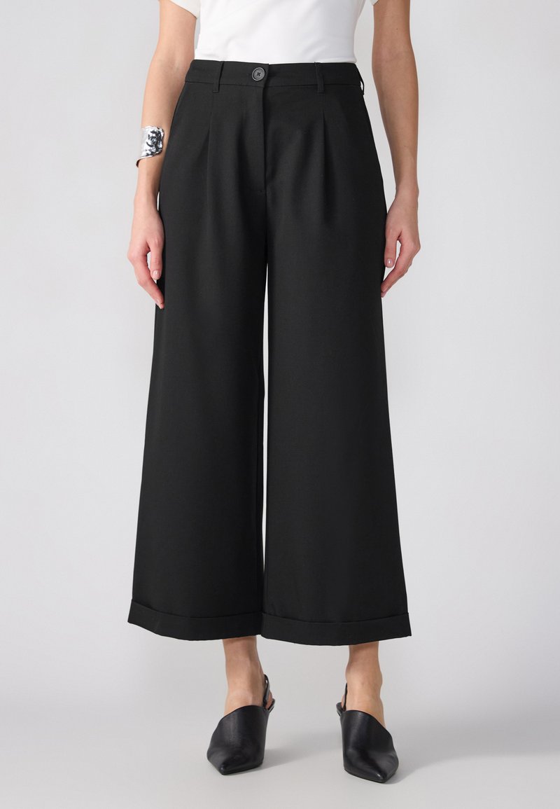 Zwarte broek met wijde pijpen en een getailleerd ontwerp, voorzien van voorplooien en een knoopsluiting. In combinatie met zwarte hakschoenen.