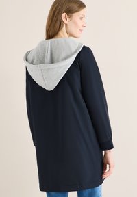 Femme portant un long manteau noir avec une grande capuche grise, vue de dos, sur un fond clair uni.