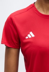 Άτομο που φοράει κόκκινη αθλητική μπλούζα με λευκό λογότυπο Adidas στο στήθος, εμφανιζόμενο από τον ώμο έως το μέσο του κορμού.