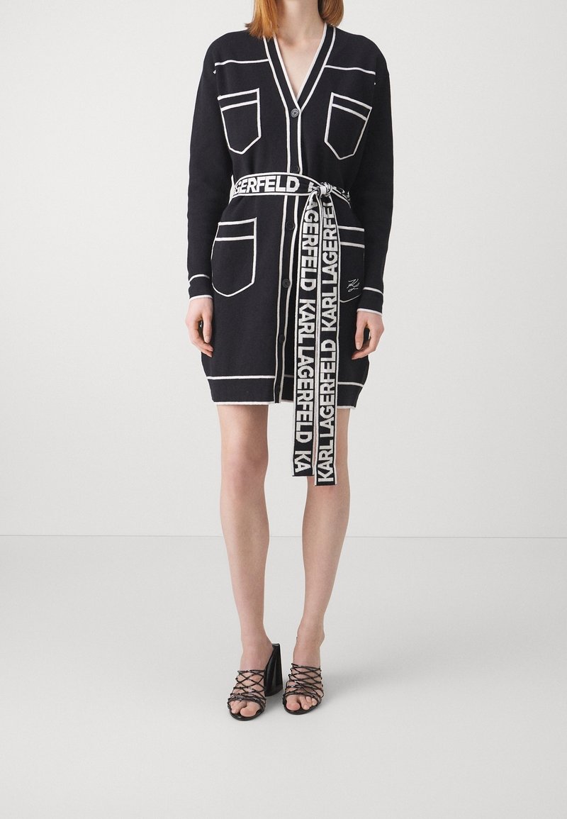 Marineblå cardigan-kjole med hvid piping, lommer og et branded bælte. Har V-hals og knaplukning. Kombineret med sorte rem-sandaler.