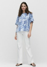Lys blå bluse med et blomstermønster, korte ærmer og rund halsudskæring, parret med hvide lige bukser og sorte sandaler.