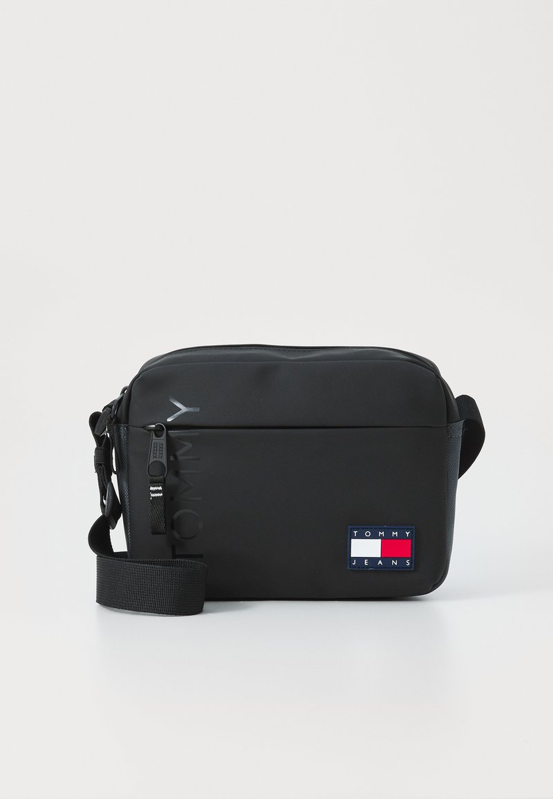 Čierna crossbody taška s hladkou textúrou, pričom sa vyznačuje výrazným logom "TOMMY" a obdlžníkovou nášivkou "TOMMY JEANS" v modrých, bielych a červených akcentoch.