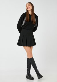 Sudadera negra con flecos plateados, acompañada de una falda negra con pliegues y borde de encaje. Llevado con botas negras hasta la rodilla. Fondo liso.