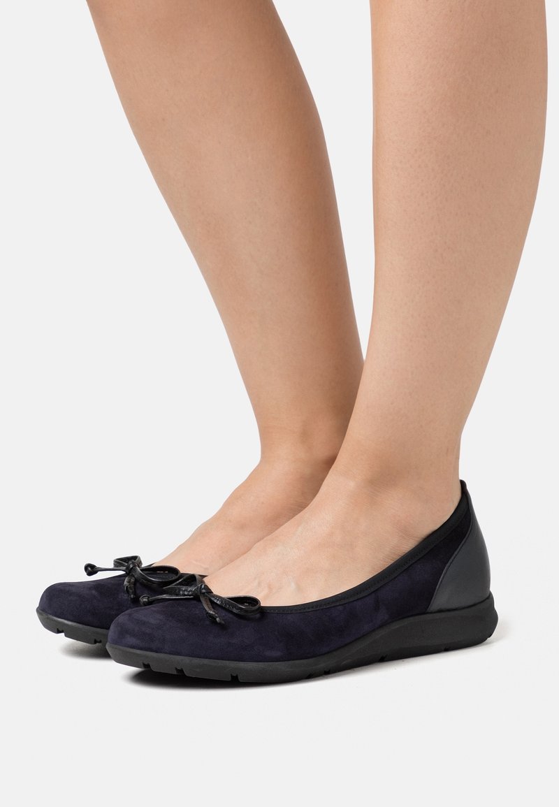 Pieds portant des ballerines en daim noir avec de petits nœuds, vus de profil sur un fond blanc uni.