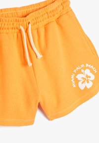 Orange, ribbede stofshorts med elastisk talje og hvid snor. Har hvid tekst "Miami Palm Beach" og hibiscusdesign på forsiden.