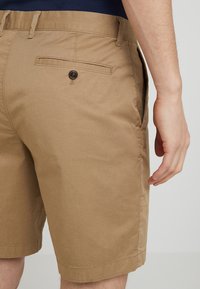 Shorts en coton kaki avec une coupe plate, des poches latérales et une poche arrière avec un bouton marron. Texture lisse et design ajusté.
