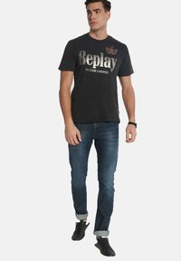 Camiseta negra con el texto plateado "Replay" y "RIDE HARD", combinada con unos vaqueros azules y zapatillas negras con los puños remangados.