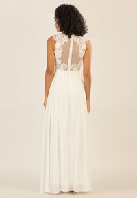 Langes weißes Kleid mit transparentem Rücken und floralem Spitzenakzenten, zarten Details am Ausschnitt und einem fließenden Chiffonrock.