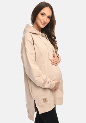 Pregnant woman with long brown hair wearing a beige hoodie and black leggings, gently holding her belly and smiling softly.